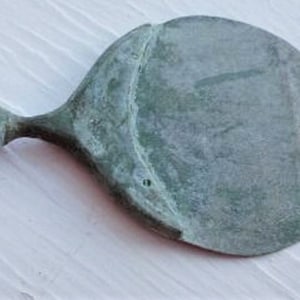 May include: An antique hand mirror with a tarnished, greenish-bronze finish. The mirror has a round face and a decorative handle. The handle has a bulbous end and a slender, hourglass-shaped midsection. The mirror is likely made of metal.