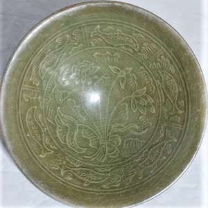May include: A celadon-colored ceramic bowl with intricate relief carvings. The bowl features a central floral design surrounded by fish and bird motifs. The rim has a slightly darker hue, adding depth to the antique aesthetic.