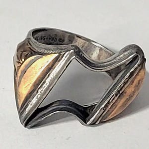 Puede incluir: Un anillo de plata con un diseño de incrustaciones de oro. El anillo está abierto en el centro y tiene una forma ondulada y orgánica.