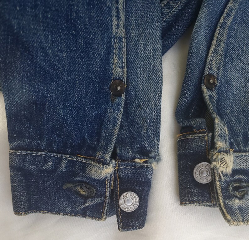 May include: Close-up of the cuffs of a blue denim jacket with metal rivets and a faded look. The cuffs are frayed and worn, showing signs of age and use.