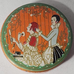 May include: Round, vintage-style decorative item with an illustration of two figures. One figure is applying makeup while the other adjusts their hair. The background is orange with floral accents, and the border is green.