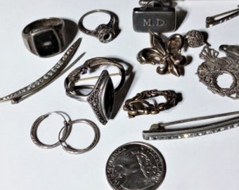 Puede incluir: Una colección de joyas y accesorios antiguos, que incluye anillos, broches, una moneda y pendientes de aro. Los artículos son de color plateado y presentan varios diseños y adornos. Un gemelo rectangular está grabado con las iniciales "M.D."