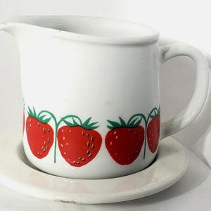 May include: A white ceramic creamer pitcher and matching saucer set featuring a repeating pattern of bright red strawberries with green stems and leaves encircling the base of the pitcher. The pitcher has a curved handle and a pouring spout.