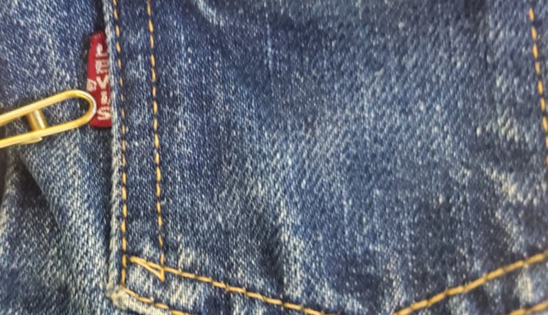 May include: Close-up of a blue denim fabric with a red Levi's label and a gold paperclip attached to it.