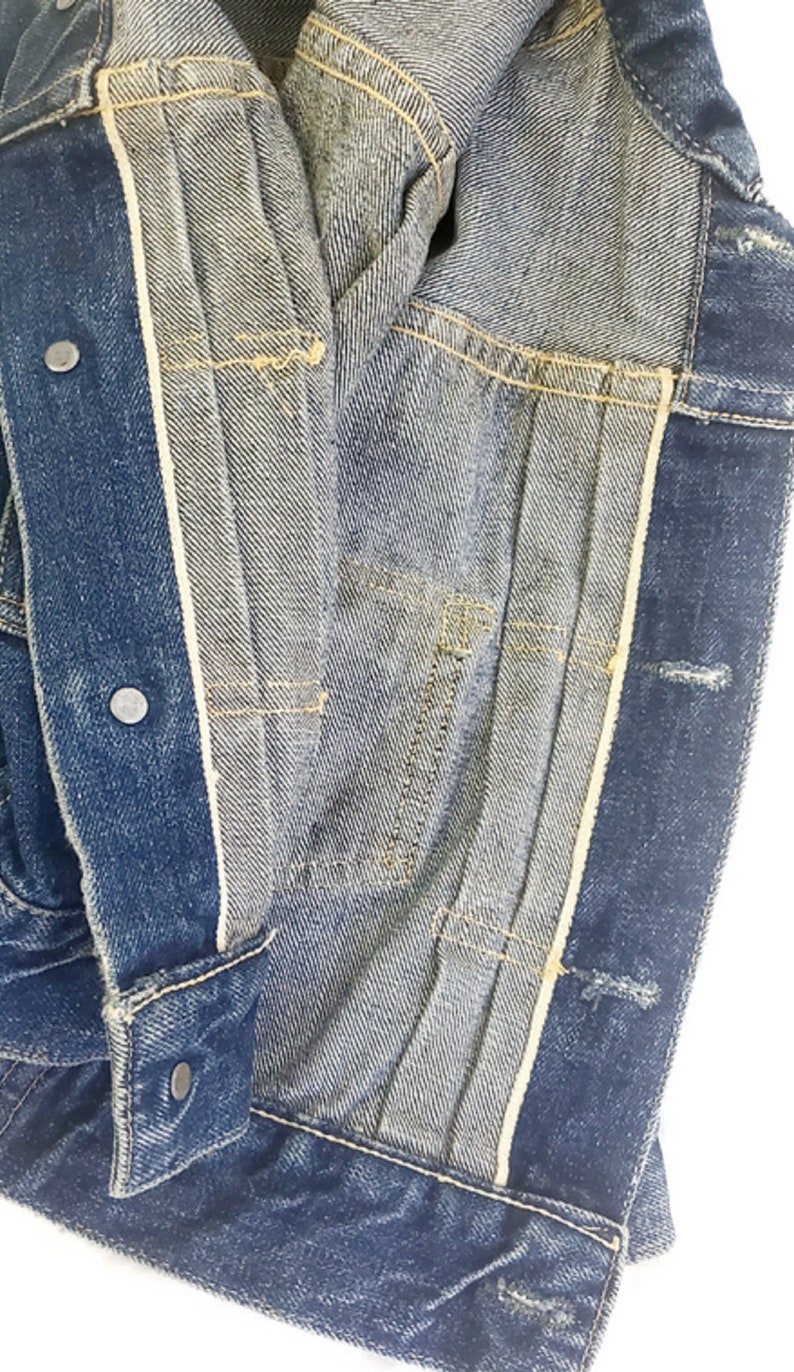 May include: Close-up of the inside of a blue denim jacket, showing the lining and stitching. The jacket is unbuttoned and the lining is visible.