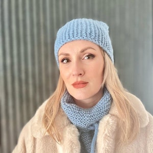Può includere: Una donna indossa un cappello e una sciarpa di lana blu chiaro. Il cappello e la sciarpa sono entrambi a coste e hanno un fiocco. Indossa un cappotto di pelliccia sintetica bianco.