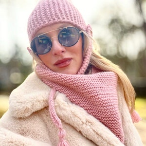 Può includere: Un set di cappello e sciarpa di lana rosa con un pon pon. Il cappello ha una patta pieghevole che copre le orecchie. La sciarpa è di forma triangolare con un lungo pon pon.