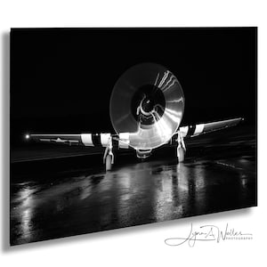 Puede incluir: Fotografía en blanco y negro de un avión antiguo, visto de frente. La hélice está enfocada, con las alas y el tren de aterrizaje visibles. La imagen está ambientada sobre un fondo oscuro, con reflejos en el suelo mojado.