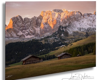 Könnte beinhalten: Eine Landschaftsaufnahme mit schneebedeckten Berggipfeln, die vom goldenen Licht des Sonnenaufgangs beleuchtet werden, über einem dunkelgrünen Nadelwald und einer Wiese mit zwei kleinen Holzhütten. Der Text Lynn A. Welles Photography ist zu sehen.