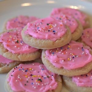 May include: A plate of pink frosted sugar cookies with colorful sprinkles. The cookies are stacked on top of each other.
