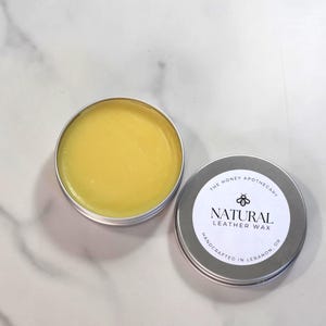 May include: Open tin of yellow leather wax next to a closed tin with a white label. The label reads "NATURAL LEATHER WAX" with a bee graphic and "THE HONEY APOTHECARY" above. The wax is handcrafted in Lebanon, OR.