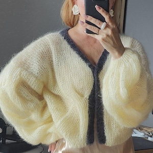 May include: A pale yellow, fuzzy, long-sleeved cardigan with a V-neckline and a dark gray trim along the front opening. The knit garment features a loose, airy texture with voluminous sleeves and a cropped length, showcasing a soft, brushed appearance.