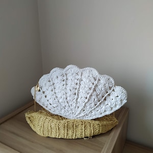 May include: A white crocheted seashell-shaped bag with a gold chain strap. The bag is sitting on a wooden surface.