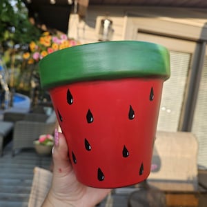 May include: A red ceramic flower pot with a green rim, painted to resemble a watermelon. Black teardrop shapes are scattered across the red surface, mimicking watermelon seeds. The pot is designed for planting flowers or herbs.