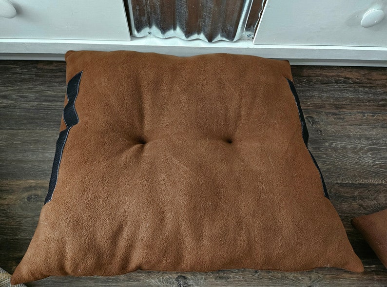 May include: A brown, rectangular floor cushion with black trim. The cushion has two buttons on the top.
