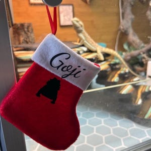 May include: Red and white Christmas stocking with the name "Goji" written in black cursive on the white cuff. A black silhouette of a lizard is embroidered on the stocking.