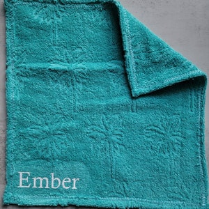 Op de afbeelding: Een turquoise keukendoek met het woord "Ember" in wit geborduurd.