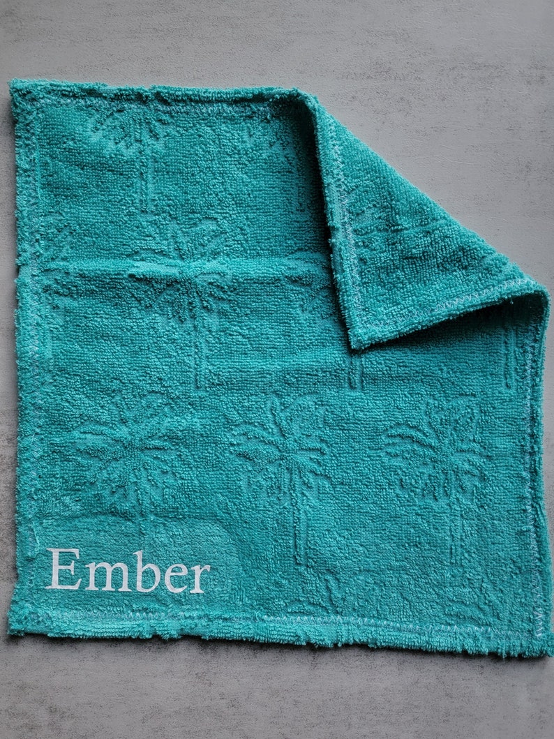 Op de afbeelding: Een turquoise keukendoek met het woord "Ember" in wit geborduurd.