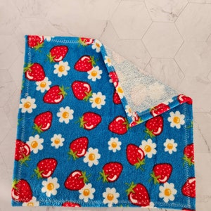Op de afbeelding: Een blauwe keukendoek met een patroon van rode aardbeien en witte madeliefjes. De handdoek is dubbelgevouwen en heeft een witte badstof achterkant.
