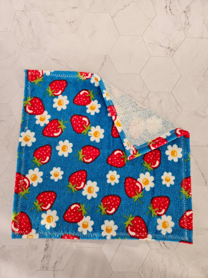 Op de afbeelding: Een blauwe keukendoek met een patroon van rode aardbeien en witte madeliefjes. De handdoek is dubbelgevouwen en heeft een witte badstof achterkant.