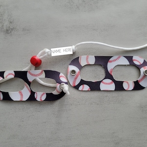 May include: Two black fabric keychains with white baseball designs and red stitching. Each keychain has a metal ring and a white cord with a red bead. One keychain has a white label with the text "NAME HERE".