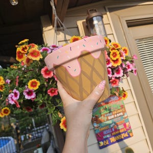 May include: A decorative ice cream cone-shaped flower pot. The pot is tan with a waffle pattern and a pink top with sprinkles and drips. The pot is held in front of a hanging basket of colorful flowers.