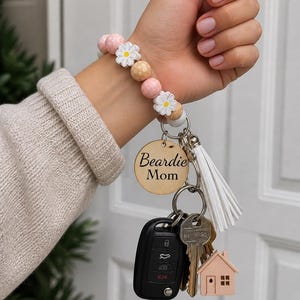 May include: A beaded keychain wristlet with a wooden tag that says "Beardie Mom". The wristlet features pink, white, and tan beads, with daisy accents. Attached are keys, a car key fob, and a small house charm.