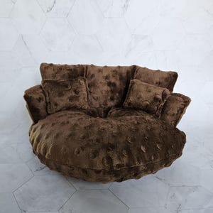 May include: A round, dark brown plush chair featuring a soft, textured fabric with a dimpled, dot-like pattern. The chair includes a large, circular base cushion and several attached back and side pillows, all covered in the same fuzzy material.