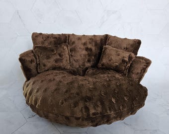 May include: A round, dark brown plush chair featuring a textured dimpled surface. The chair includes a large, circular base cushion and a curved backrest section, accompanied by two matching square throw pillows placed on the seat.