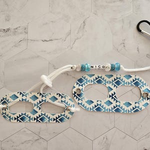 May include: Two white and blue patterned fabric face mask holders with a white cord and a beaded name tag that says "NICO".