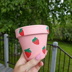 May include: A pink terracotta flower pot decorated with red strawberries and green leaves. The pot is held in a hand, with a blurred green background of trees and grass.