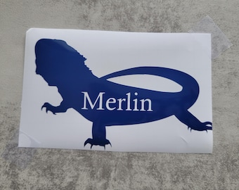 May include: A white sticker with a blue silhouette of a bearded dragon with the name "Merlin" written in white text.