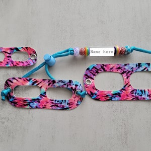 May include: A colorful tie-dye patterned lanyard with a blue cord and a name tag that says "Name here". The lanyard has two loops for attaching to a badge or ID card.