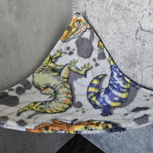 K&ouml;nnte beinhalten: Eine dreieckige Haustierh&auml;ngematte aus weichem, wei&szlig;em Fleece-Stoff mit einem bunten Dinosaurier-Aufdruck, der gelbe, gr&uuml;ne, blaue und orangefarbene Reptilien zeigt. Die H&auml;ngematte verf&uuml;gt &uuml;ber Metall&ouml;sen an jeder Ecke zur sicheren Befestigung.
