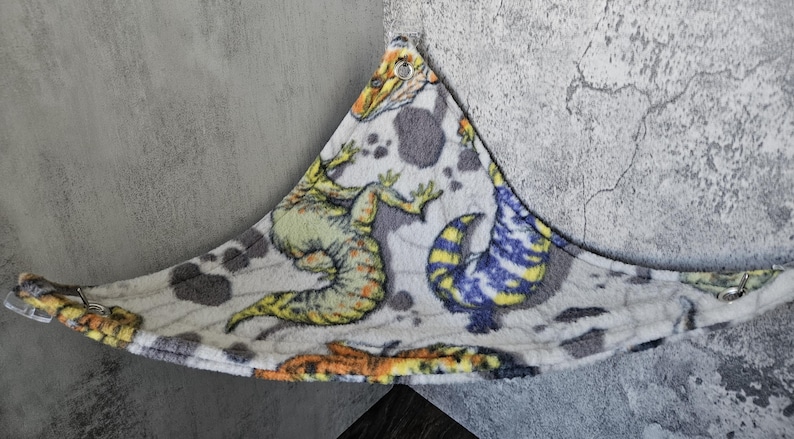 K&ouml;nnte beinhalten: Eine dreieckige Haustierh&auml;ngematte aus weichem, wei&szlig;em Fleece-Stoff mit einem bunten Dinosaurier-Aufdruck, der gelbe, gr&uuml;ne, blaue und orangefarbene Reptilien zeigt. Die H&auml;ngematte verf&uuml;gt &uuml;ber Metall&ouml;sen an jeder Ecke zur sicheren Befestigung.