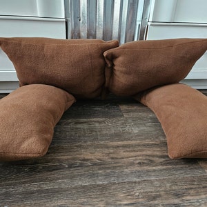 May include: Two brown fleece pillows are arranged in a U-shape on a wooden floor. The pillows are soft and plush, and they would be perfect for lounging or reading.