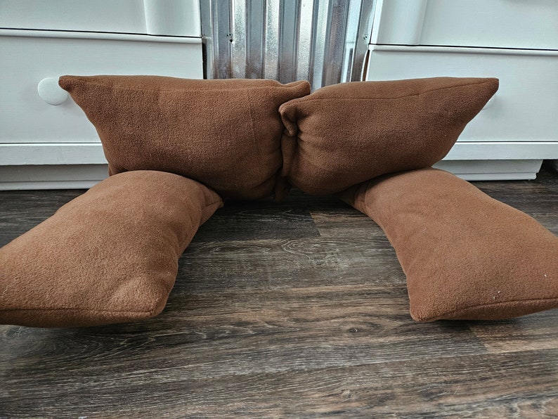 May include: Two brown fleece pillows are arranged in a U-shape on a wooden floor. The pillows are soft and plush, and they would be perfect for lounging or reading.