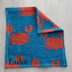 Op de afbeelding: Een blauwe en oranje handdoek met een krabbenpatroon en de naam "Luke" geborduurd.