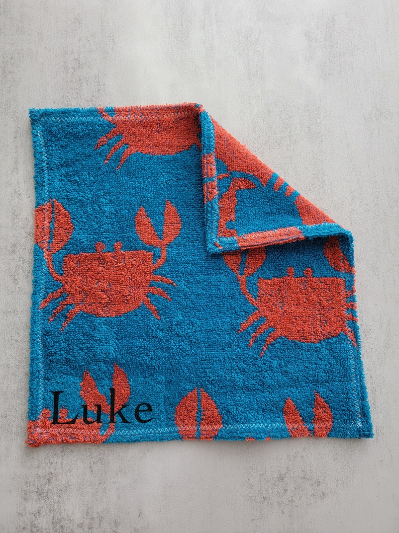 Op de afbeelding: Een blauwe en oranje handdoek met een krabbenpatroon en de naam "Luke" geborduurd.