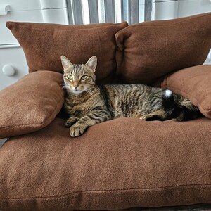 May include: A brown cat with tabby markings is lying on a brown plush cat bed. The bed has multiple cushions and is designed for comfort.