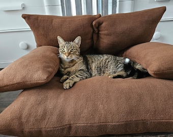 May include: A brown cat with tabby markings is lying on a brown plush cat bed. The bed has multiple cushions and is designed for comfort.