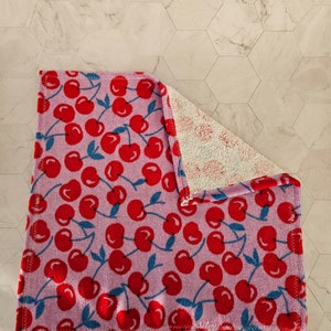 Op de afbeelding: Een roze keukendoek met een rode kersenprint. De handdoek is dubbelgevouwen en heeft een witte badstofkant.