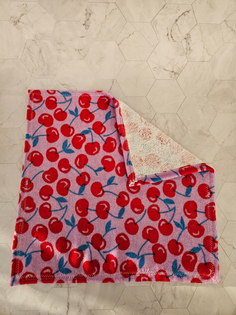 Op de afbeelding: Een roze keukendoek met een rode kersenprint. De handdoek is dubbelgevouwen en heeft een witte badstofkant.