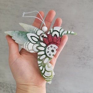 May include: A green and white hummingbird ornament with a red and white mandala design. The ornament has a white bead at the top for hanging.