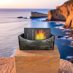 Puede incluir: Un porta tarjetas de madera tallado con una tortuga y flores de hibisco. El porta tarjetas está sobre una superficie de piedra con un libro titulado "Poetry Inspired From" de Kellie Earnshaw en el fondo.