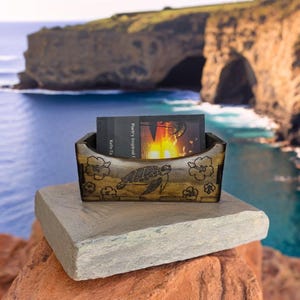 Puede incluir: Un porta tarjetas de madera tallado con una tortuga marina y flores de hibisco. El porta tarjetas está colocado sobre una roca gris con una vista de una costa rocosa y el océano en el fondo. El porta tarjetas contiene una tarjeta de visita que dice "Poetry Inspired by Killie Fa."