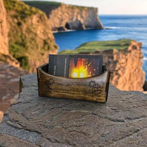 Puede incluir: Un porta tarjetas de madera con un diseño tallado de palmeras. El porta tarjetas está lleno de una tarjeta de visita que dice "Poetry Inspired From" y "Kellie Farr". El porta tarjetas está sentado en una roca con vista a una costa rocosa y el océano.