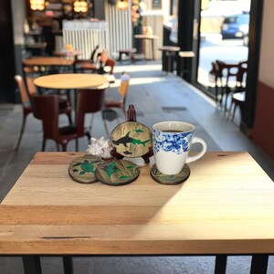 Op de afbeelding: Houten onderzetters met zeedierontwerpen, waaronder een haai en een schildpad, worden getoond op een houten tafel. Een witte mok met een blauw bloemenpatroon staat op een bijpassende onderzetter. De scène speelt zich af in een café.