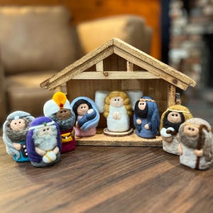 May include: A felt nativity set featuring a wooden stable structure and several small figures, including an angel with golden hair, a central family unit, and various robed characters in shades of blue, yellow, and brown.