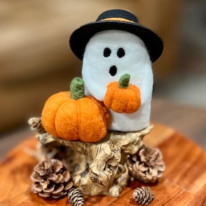 May include: A white felt ghost figurine wearing a black hat with an orange band, holding a small orange pumpkin. A larger orange pumpkin sits beside the ghost on a textured wooden base decorated with several brown pinecones.
